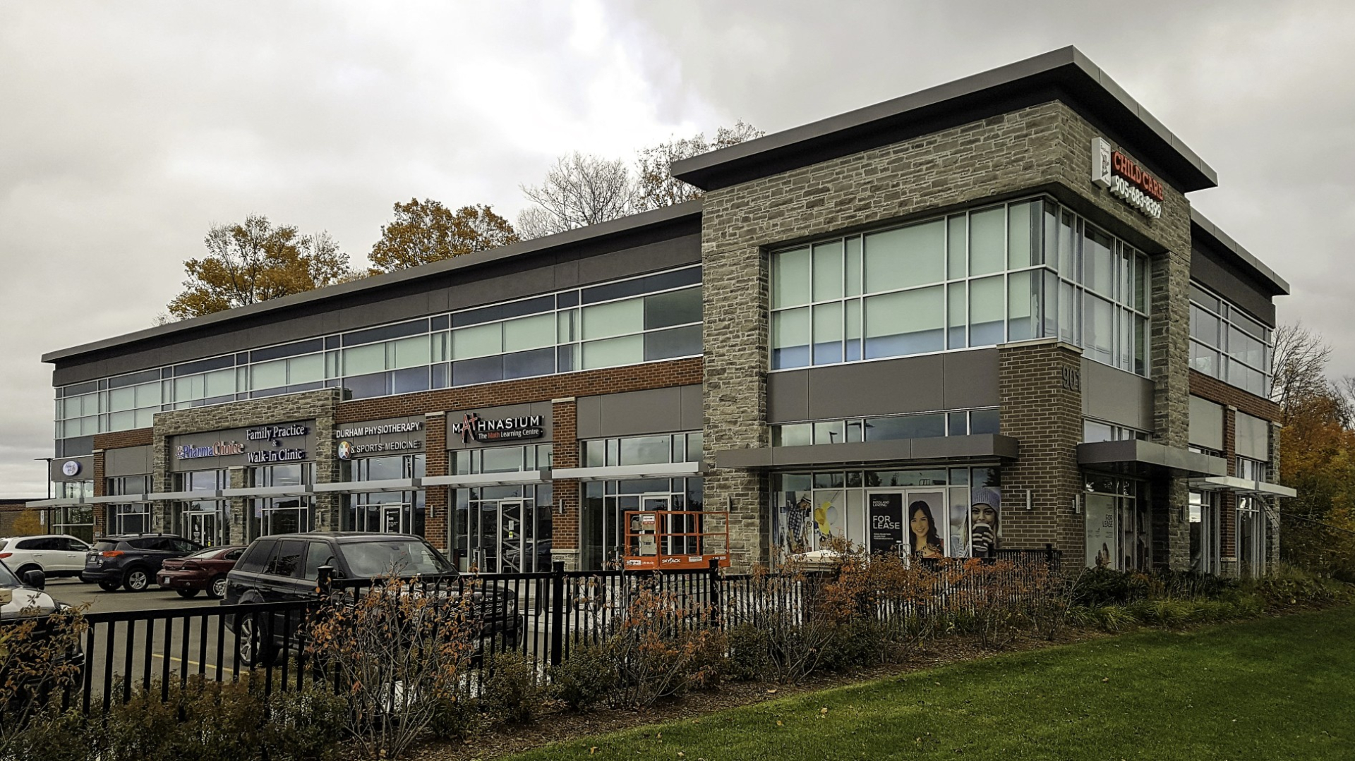 A modern two‑storey retail plaza with large glass windows, brick and stone exterior walls, and multiple storefront entrances. The building is bordered by landscaping and a small parking area under an overcast sky.